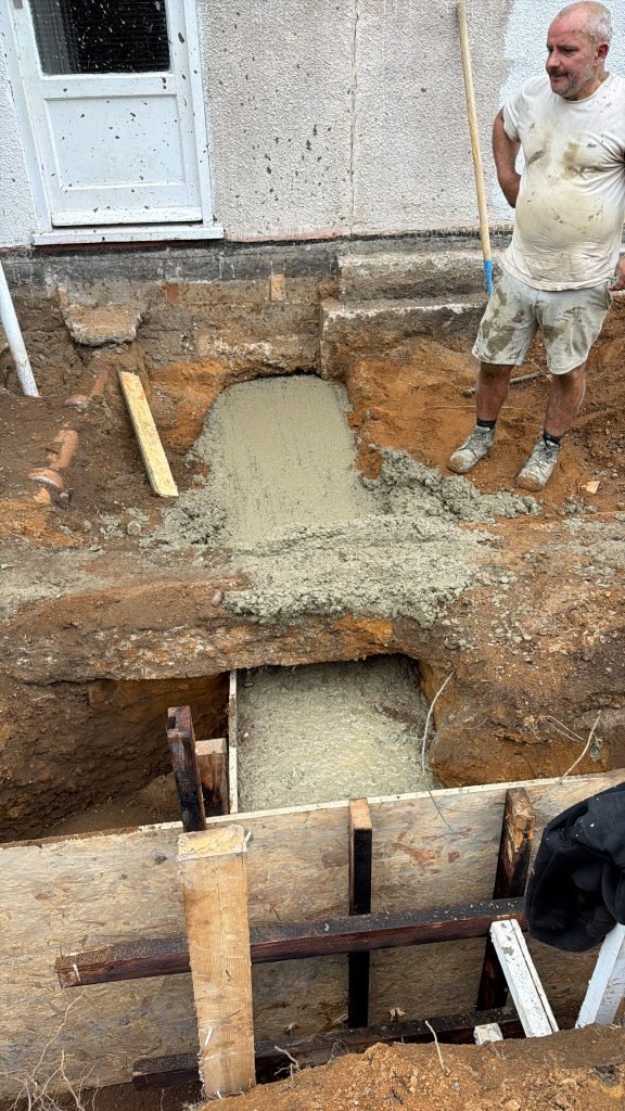 Worker stands beside a freshly poured concrete footing in a trench next to a house wall, wearing dirt-stained clothes and boots.
