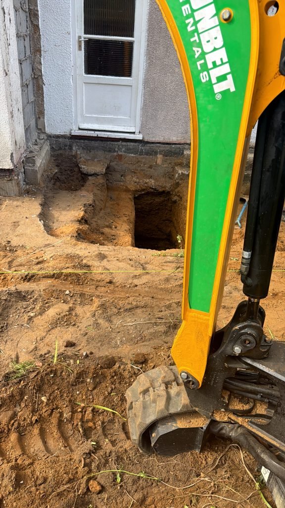 Excavation next to a house entrance with a white door, exposing soil and a trench beside the foundation, and a small green-yellow excavator in the foreground.