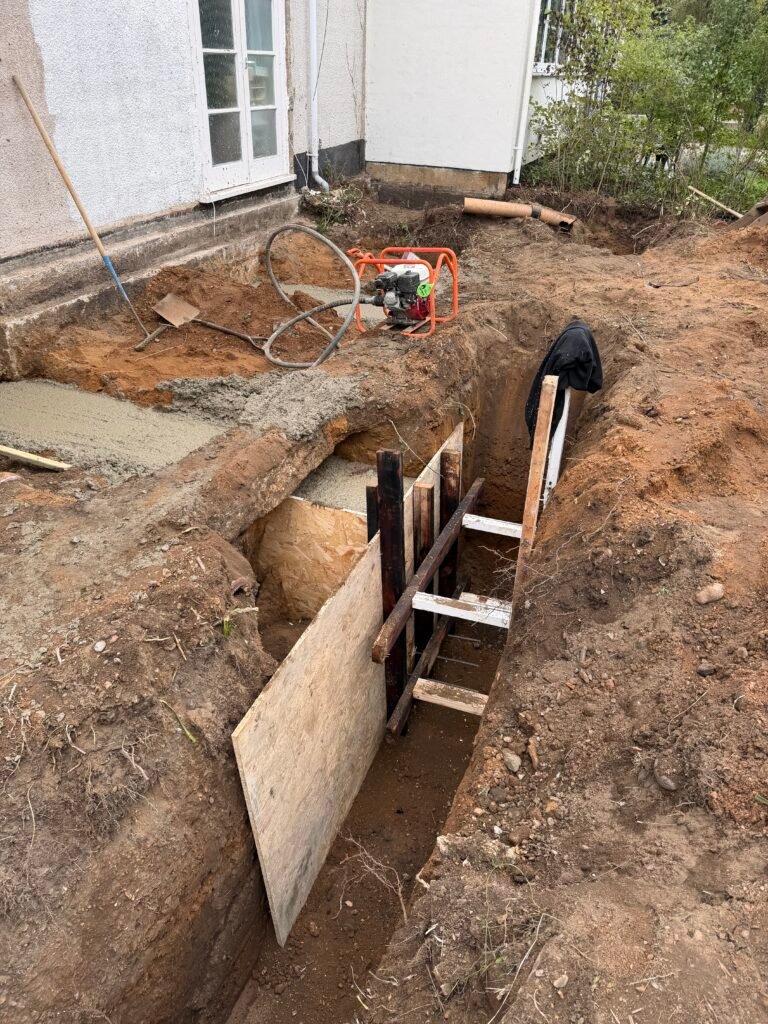 Trench beside a house foundation with wooden panels and an orange-framed portable pump in the dirt.