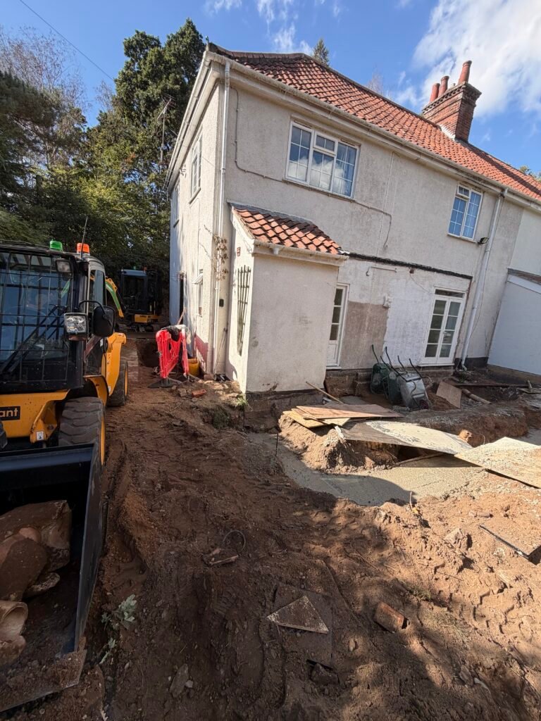 Construction site beside a white two-story house with a red-tiled roof; dirt piles and machinery in the foreground.
