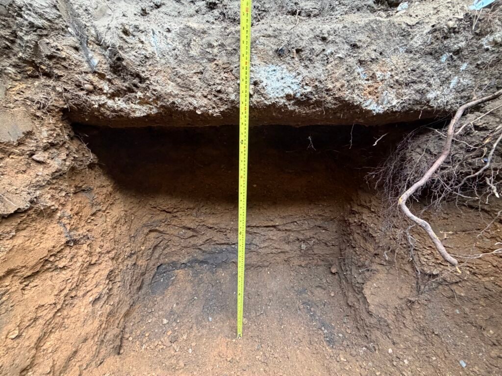 Soil trench with exposed layers and a yellow measuring tape standing upright to indicate depth in the wall of the excavation site.